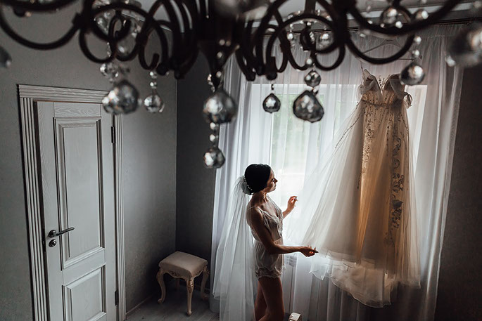 Невеста рассматривает свадебное платье у окна.